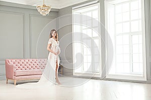 Beautiful pregnant young woman in white peignoir standing near pink sofa, holding with love her belly
