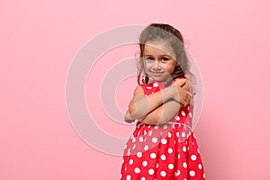 Beautiful preschool 4 years kid girl shyly hugging herself, cute smiling posing on camera against pink wall background with copy