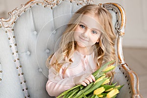 Beautiful pretty girl with yellow flowers tulips sitting in arm-chair, smiling. Indoor photo