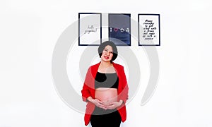Beautiful and pretty pregnant woman standing with smiling and touching or holding her belly with white wall background