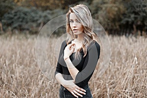 Beautiful pretty sexy young woman in a black stylish T-shirt with blond hair in trendy jeans poses in a field on a warm autumn day