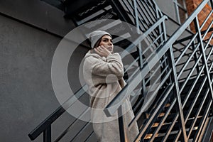 Beautiful pretty stylish young woman in a fashionable knitted hat in a trendy milk-colored faux-fur coat posing on a metallic