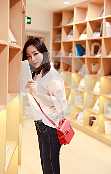 A beautiful pretty young girl woman female carry a small red bag read book at a book store library model look at camera