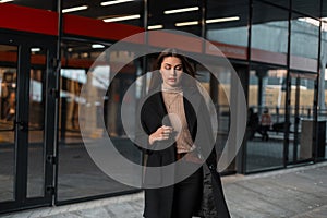 Beautiful pretty young woman with brown hair in a long black elegant coat in a beige vintage shirt stands on the street.