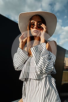 Beautiful pretty young woman in fashion striped dress in stylish sunglasses in vintage straw hat posing outdoors on bright sunny