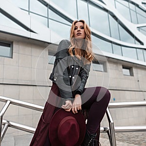 Beautiful pretty young woman in a fashionable leather jacket in purple vintage pants with a hat posing near a modern building