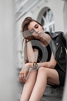 Beautiful pretty young woman in a fashionable short black dress in a stylish leather black jacket is resting on the staircase in