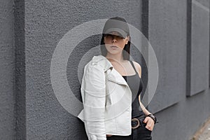 Beautiful pretty young woman in a stylish black baseball cap in a fashionable white leather jacket in vintage jeans in a t-shirt