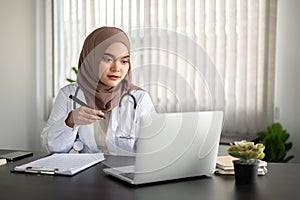 A beautiful Asian Muslim female doctor is working on her laptop at her desk at a hospital or clinic