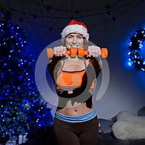 Beautiful red-haired female athlete in a Santa Claus hat posing in a dark studio. Fitness woman in sportswear holds a