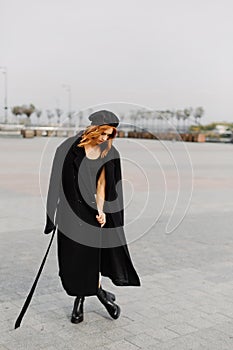 Beautiful red-haired girl in a black coat and a beret walks through the city