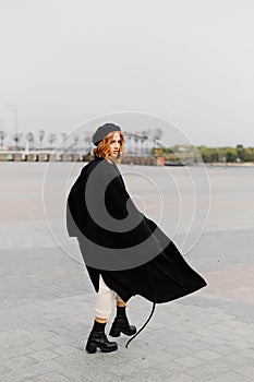 Beautiful red-haired girl in a black coat and a beret walks through the city