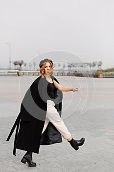 Beautiful red-haired girl in a black coat and a beret walks through the city