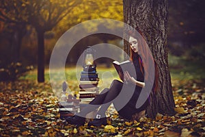 Beautiful red-haired girl with books and a lantern sitting under a tree in the autumn fairy forest. A fabulous autumn
