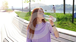 Beautiful red-haired girl drinking water from bottle sitting on bench in park on bright sunny day.