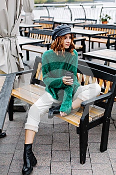 Beautiful red-haired girl in a green sweater and cap sits with a cup of coffee in a cafe on the street