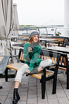 Beautiful red-haired girl in a green sweater and cap sits with a cup of coffee in a cafe on the street