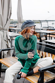 Beautiful red-haired girl in a green sweater and cap sits with a cup of coffee in a cafe on the street