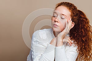 Beautiful red-haired girl with long curly hair in a white sweater is sad and looks down. Shot in a studio on a beige background