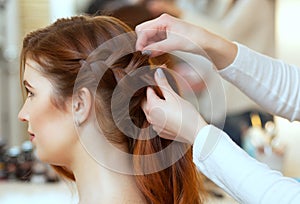 Beautiful, red-haired girl with long hair, hairdresser weaves a French braid, in a beauty salon