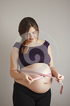 Beautiful red-haired pregnant woman use tape measure at big belly on gray studio background, female body during pregnancy