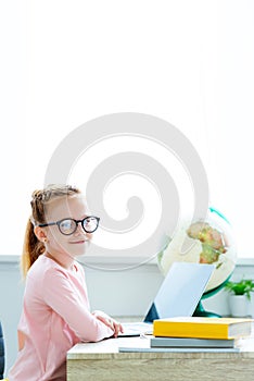 beautiful red haired schoolgirl in eyeglasses smiling at camera while studying with books and laptop