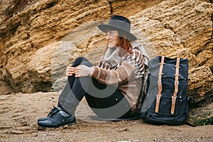 Beautiful red-haired woman in a hat and a scarf with a backpack sits on the coast on the background of the rocks. Tourism, rest,