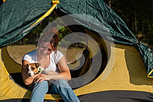 Beautiful red-haired woman is resting in nature with her pet. A girl sits in a tourist tent and hugs a Jack Russell