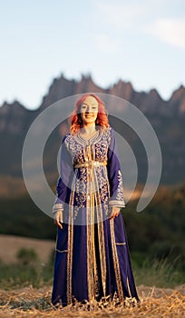 Beautiful red-haired woman in a traditional blue dress with goldwork ornamentations in a sunny field