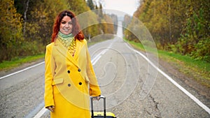 Beautiful red-haired woman in a yellow coat walks with a yellow suitcase along the highway in autumn.