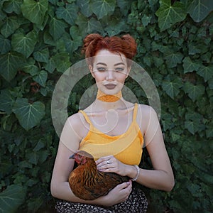 A beautiful red-haired young woman holds a brown hen in her arms against a background of emerald green ivy. Artistic