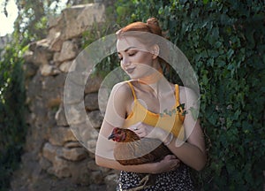 A beautiful red-haired young woman in the village holds a brown hen in her arms against a background of emerald green