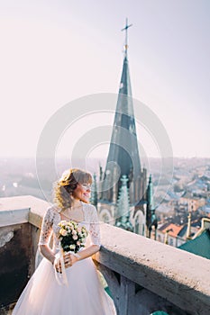 Beautiful red-head bride posing with bouquet on old castle balcony, cityscape background
