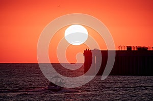 Beautiful red and orange sunset over the sea. The sun goes down over the sea. Silhouette of a pier with fishermen and boat at