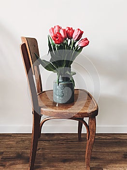 Beautiful red tulips in stylish vase on wooden rustic chair in home. Hello spring concept. Countryside living. Modern rural still