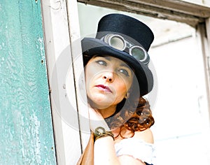Beautiful redhair steampunk girl with goggles on black hat outside toilet background. Old-fashioned