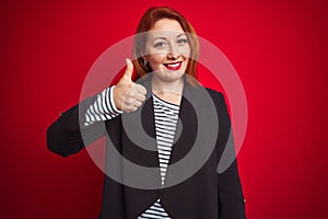 Beautiful redhead business woman wearing elegant jacket over isolated red background doing happy thumbs up gesture with hand