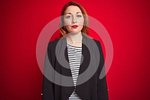 Beautiful redhead business woman wearing elegant jacket over isolated red background with serious expression on face