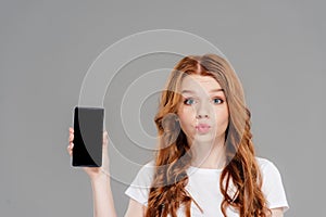 Beautiful redhead girl with duck face looking at camera and showing smartphone with blank screen isolated on grey.