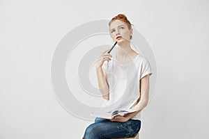 Beautiful redhead girl making notes thinking sitting over white background.