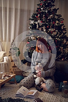 Beautiful redhead girl playing with a cat near a Christmas tree