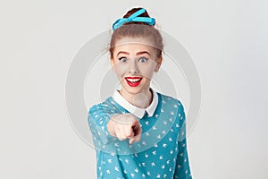 The beautiful redhead girl, wearing blue dress, opening mouths widely, having surprised shocked looks, pointing finger at camera.