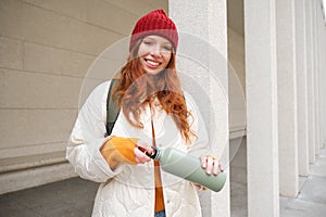 Beautiful redhead woman drinking hot tea or coffee from thermos, female tourist enjoys warm drink, rests during her