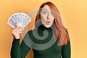 Beautiful redhead woman holding 2 singapore dollars banknotes scared and amazed with open mouth for surprise, disbelief face