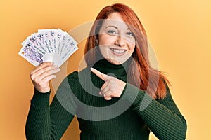 Beautiful redhead woman holding 2 singapore dollars banknotes smiling happy pointing with hand and finger