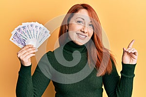 Beautiful redhead woman holding 2 singapore dollars banknotes smiling happy pointing with hand and finger to the side