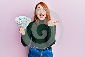 Beautiful redhead woman holding 20 hong kong dollars banknotes pointing thumb up to the side smiling happy with open mouth