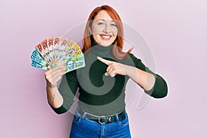 Beautiful redhead woman holding australian dollars smiling happy pointing with hand and finger