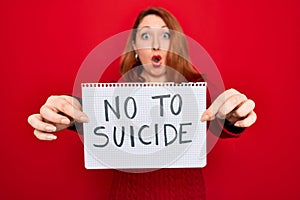 Beautiful redhead woman holding banner showing no to suicide message over red background scared in shock with a surprise face,