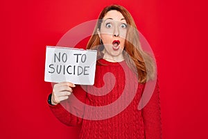 Beautiful redhead woman holding banner showing no to suicide message over red background scared in shock with a surprise face,